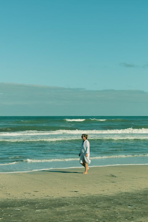 Free A woman walks along a calm beach, wrapped in a towel, with soft ocean waves in the background. Stock Photo