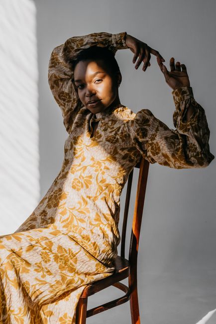 A stylish woman in a floral dress strikes a pose on a chair, enhanced by dramatic sunlight and shadow.