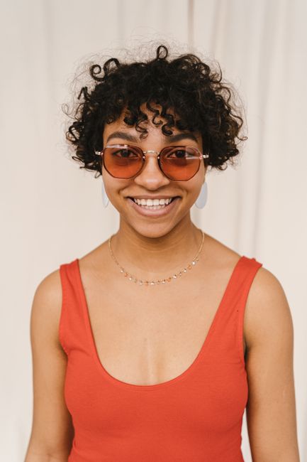 Portrait of a smiling woman with curly hair and sunglasses in an orange tank top.