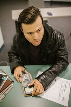 College student using a calculator and test paper, possibly during an exam or study session, in a classroom setting.