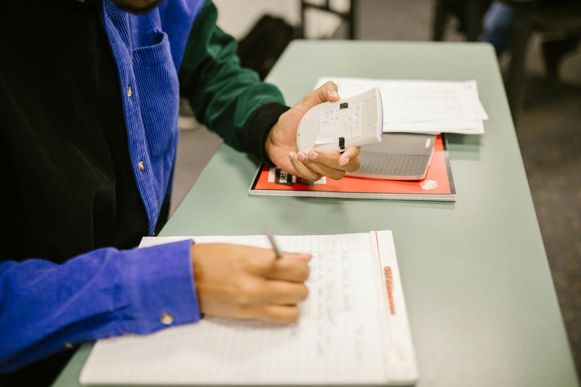 Student using a calculator and taking notes during exam preparation in a classroom.