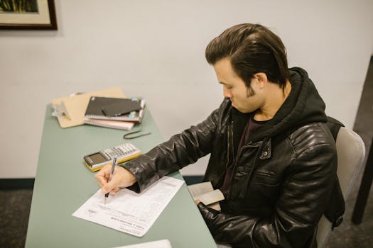 Man in leather jacket writing test answers in college classroom setting.