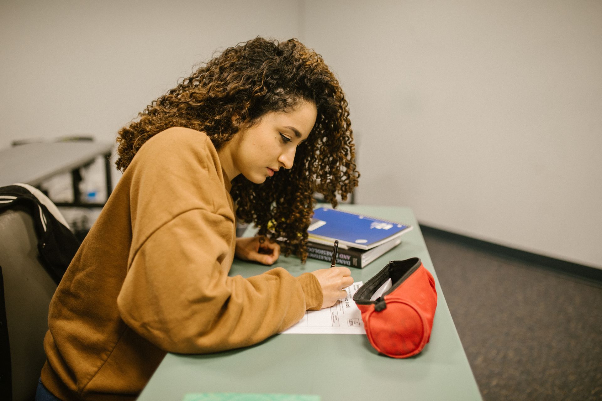 A young woman concentrating on her writing at a desk, surrounded by study materials in a classroom.