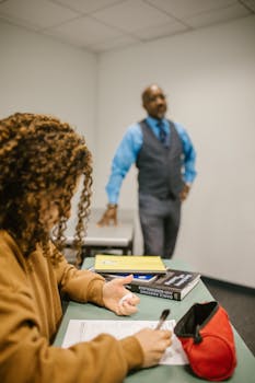 A college student writing an exam in class under the watchful eye of a professor.