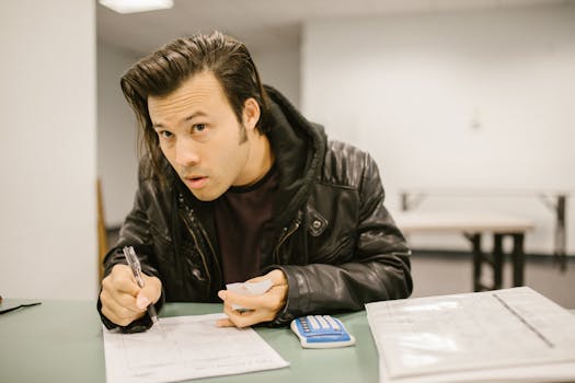 Adult male student writing in a classroom setting, holding a cheat sheet with a calculator nearby.