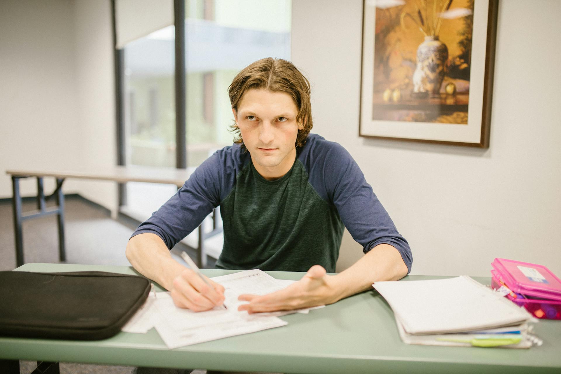 Young man looking frustrated while sitting at a desk with exam papers in a classroom.