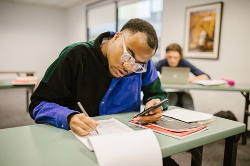 Free Student sitting at desk using smartphone while preparing for an exam in a classroom setting. Stock Photo