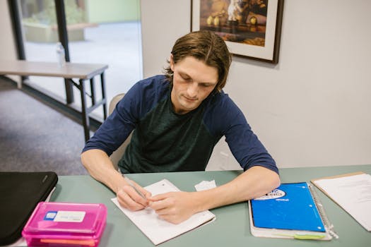 Young man writing notes at desk in a college classroom setting.