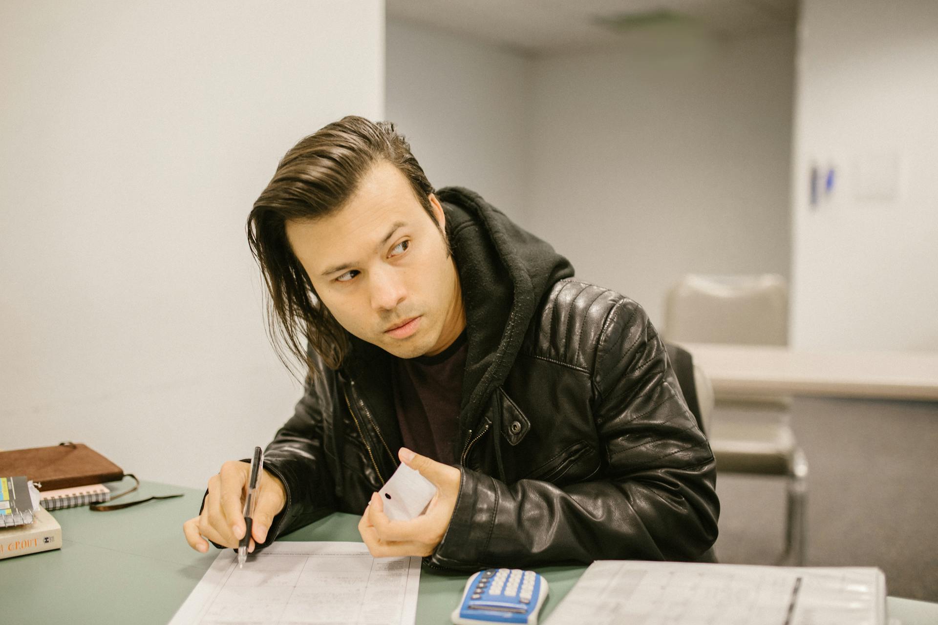 Young man in a classroom looking away while holding a phone, possibly cheating. Exam setting with calculator and paper.