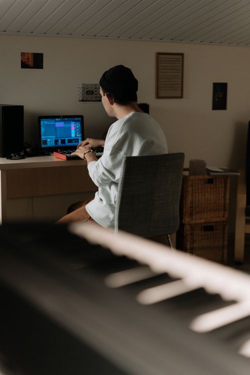 Free Back view of a man using a computer for music production, seated indoors in a modern home office. Stock Photo