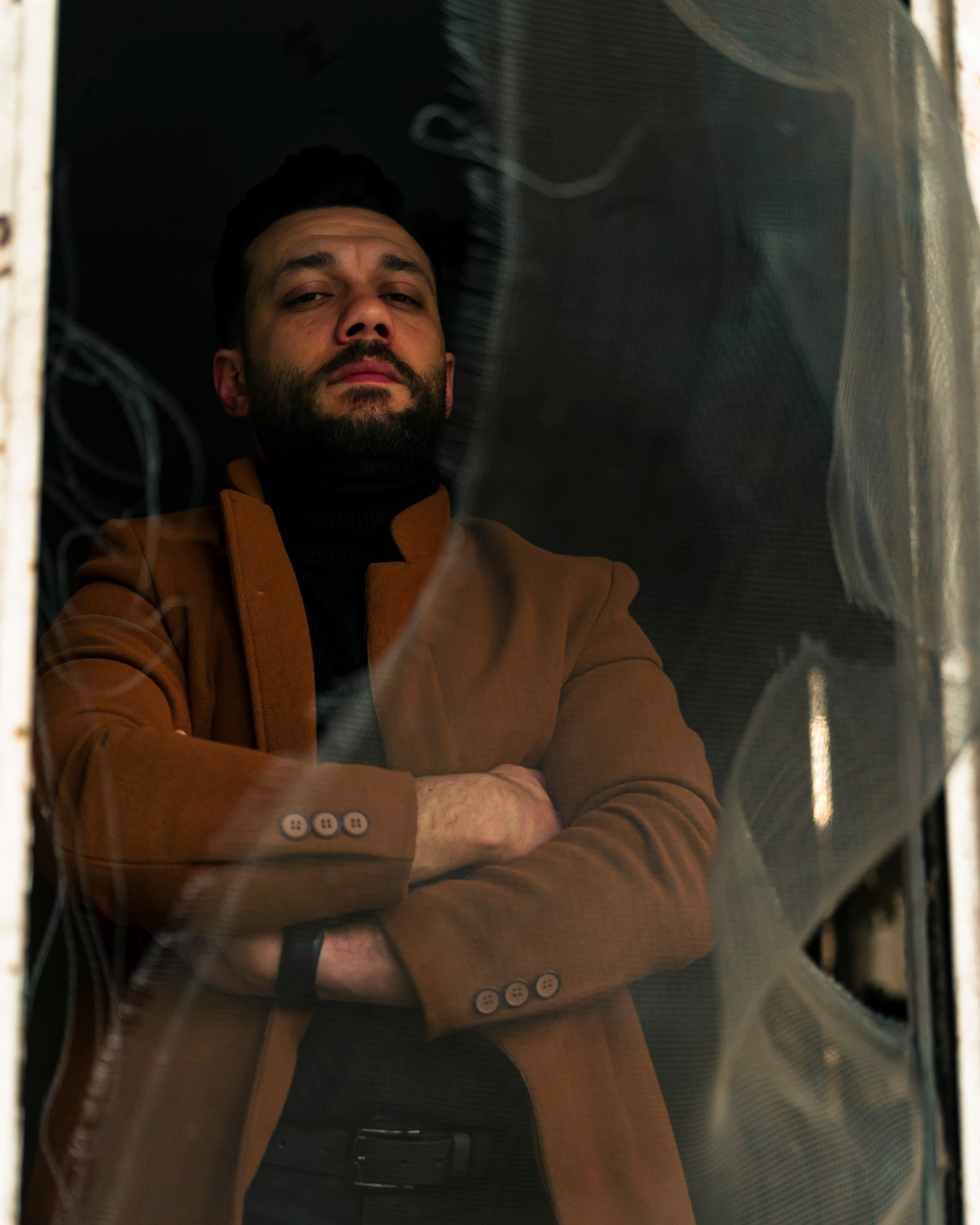 A fashionable man poses confidently with arms crossed, wearing a stylish brown coat in a low-lit interior setting.