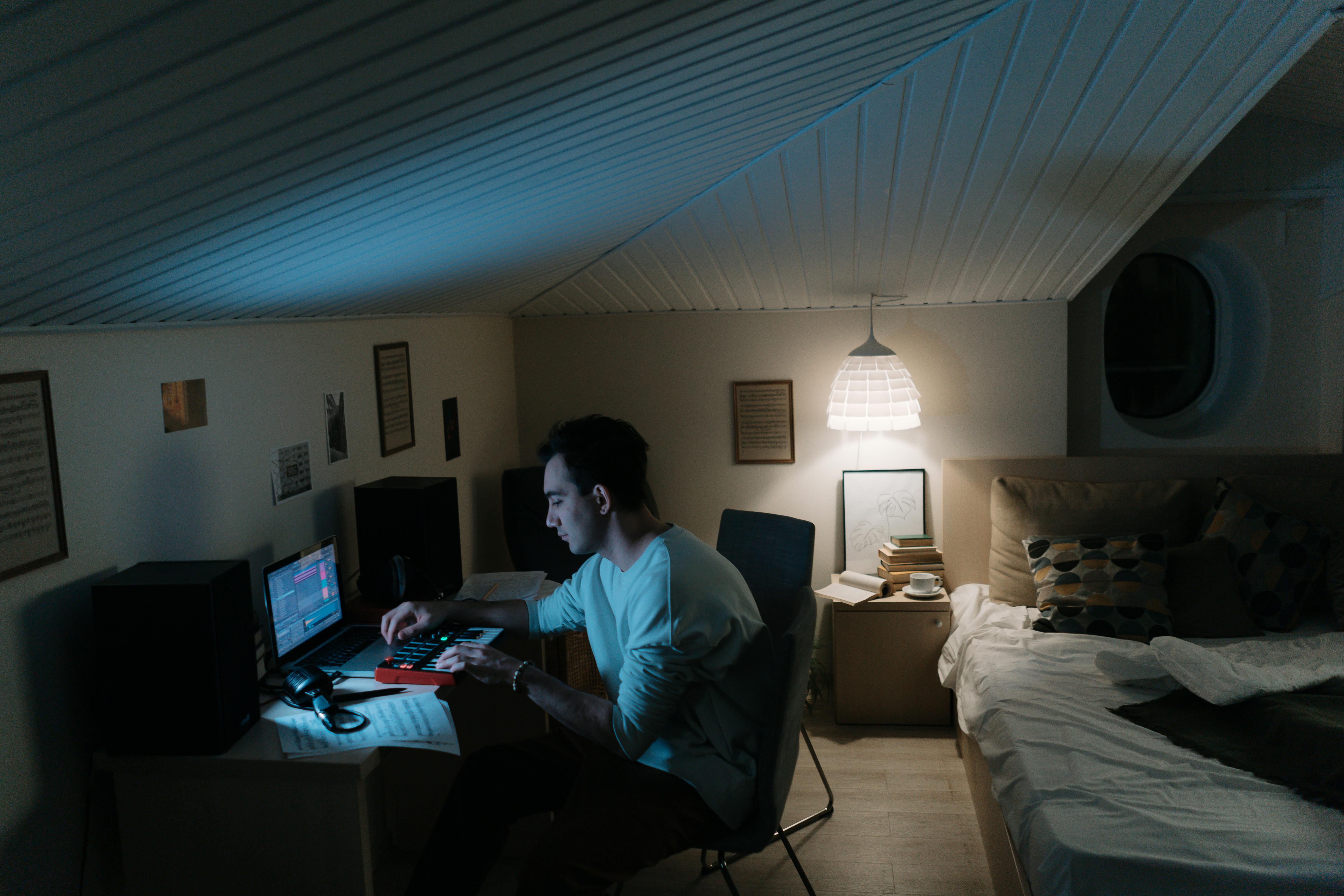 Adult man composing music in a cozy home studio, focused on a laptop and synthesizer under warm ambient lighting.