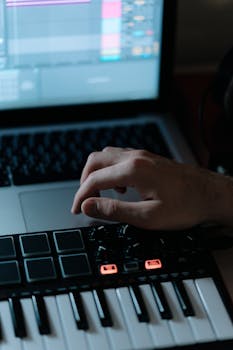 Close-up of hand adjusting synthesizer controls with laptop screen displaying audio software.