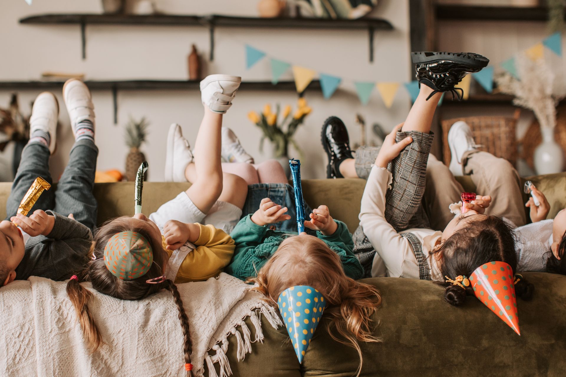 Kids lying on a sofa wearing party hats and playing with noise blowers in a festive indoor setting.