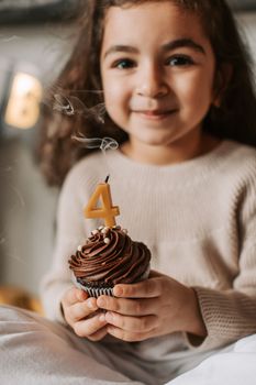 Smiling girl with a chocolate cupcake and a '4' candle, celebrating her birthday indoors.