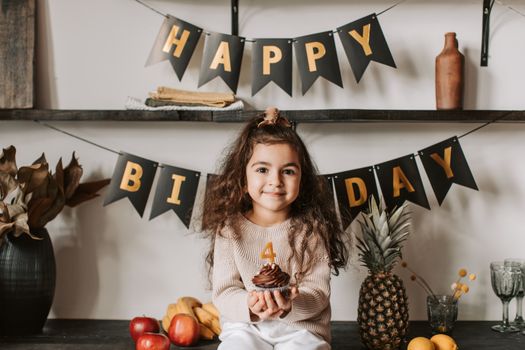 A smiling girl celebrates her birthday with a chocolate cupcake and fruit decorations indoors.