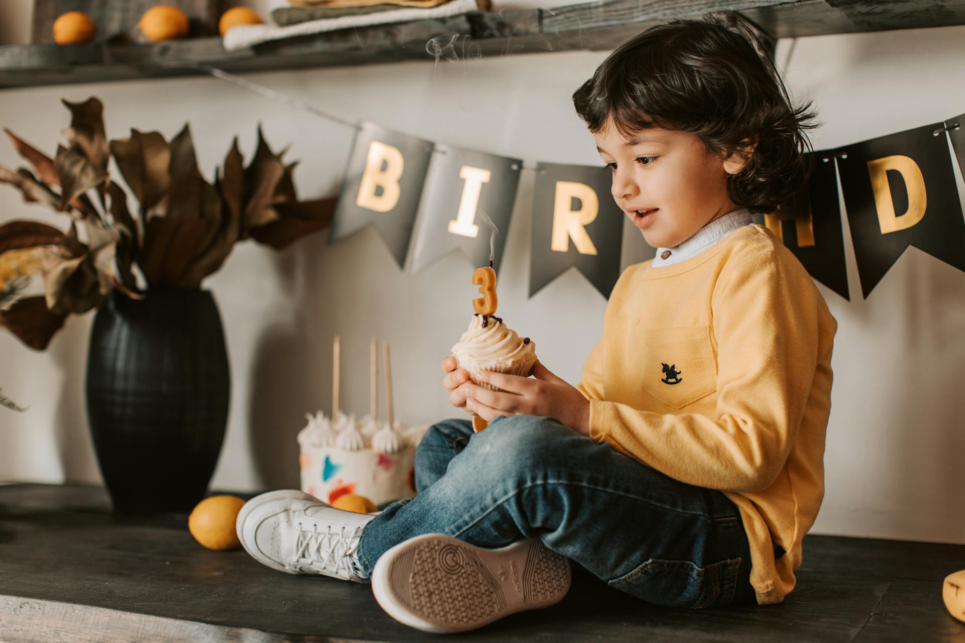 A young child celebrates a birthday indoors, holding a cupcake with a candle.