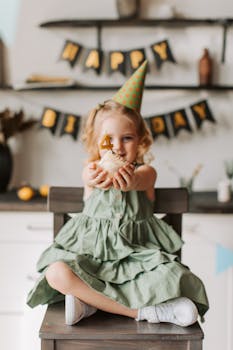 A smiling child in a green dress and party hat holding birthday cupcake indoors.