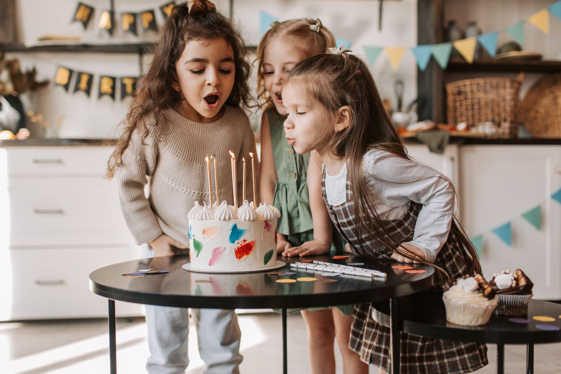 Three children blowing candles on a colorful birthday cake indoors with cheerful decorations.