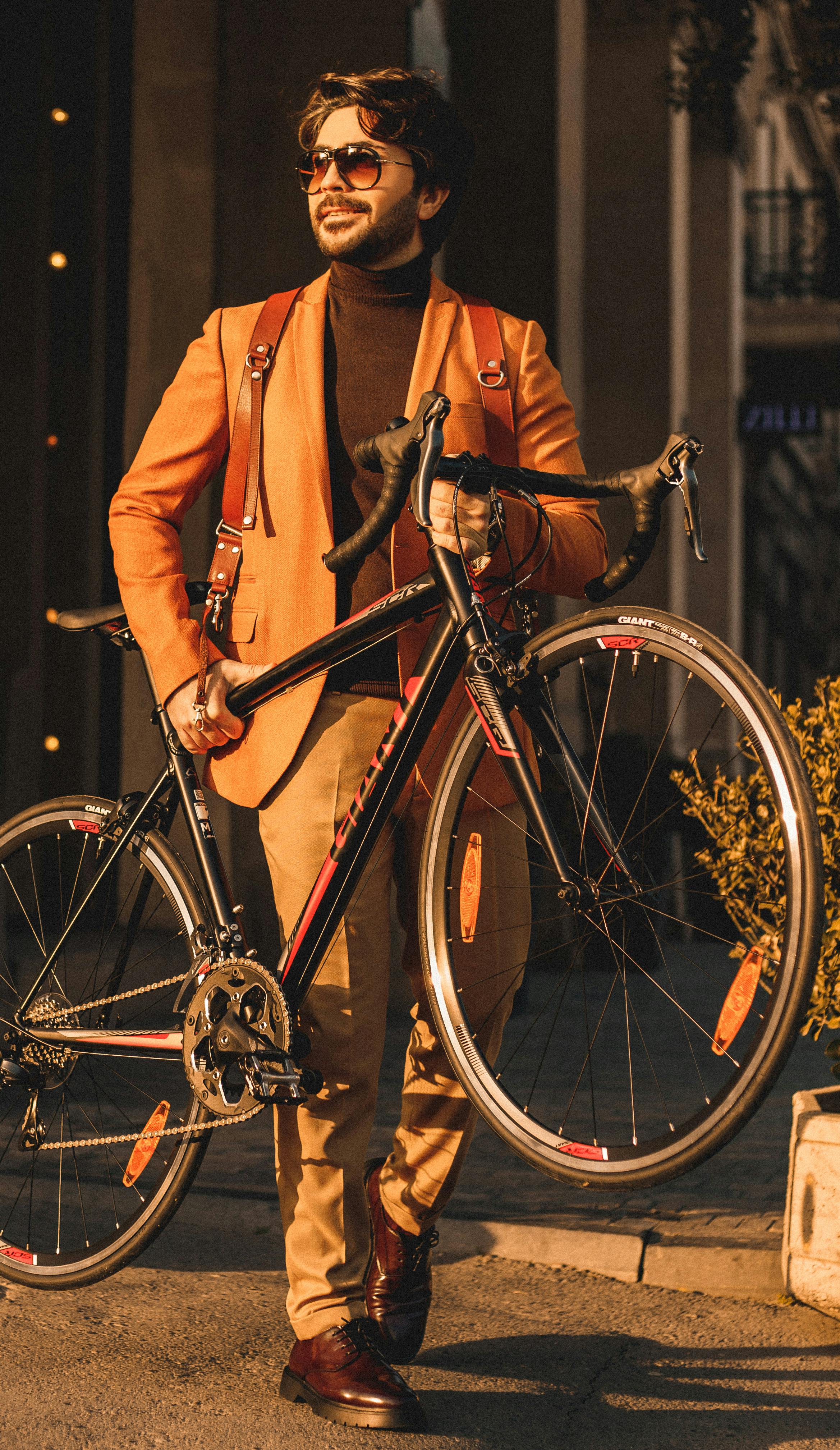 Full body of bearded male in trendy clothes walking on street with bicycle in hands on street with bright sunlight