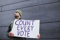 A Man in Green Beanie Holding a Placard on the Street