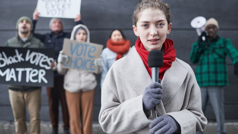 Female journalist reporting live from a protest with diverse activists and signs advocating voting rights.