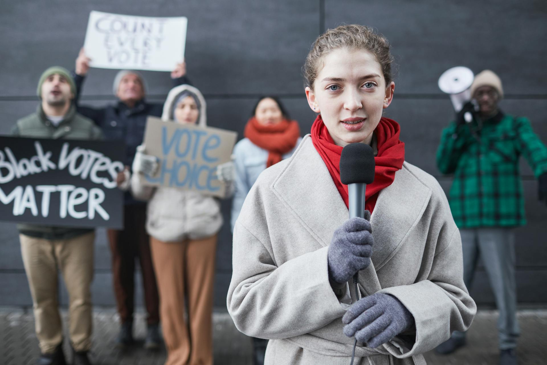 Female journalist reporting live from a protest with diverse activists and signs advocating voting rights.