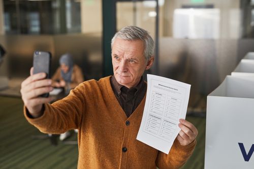 Free Elderly man with gray hair holding a ballot and taking a selfie in a voting booth. Stock Photo