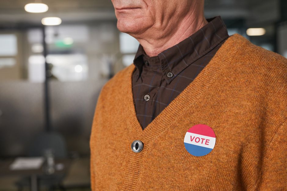 Close-up of a senior man in an orange sweater wearing a Vote badge indoors.