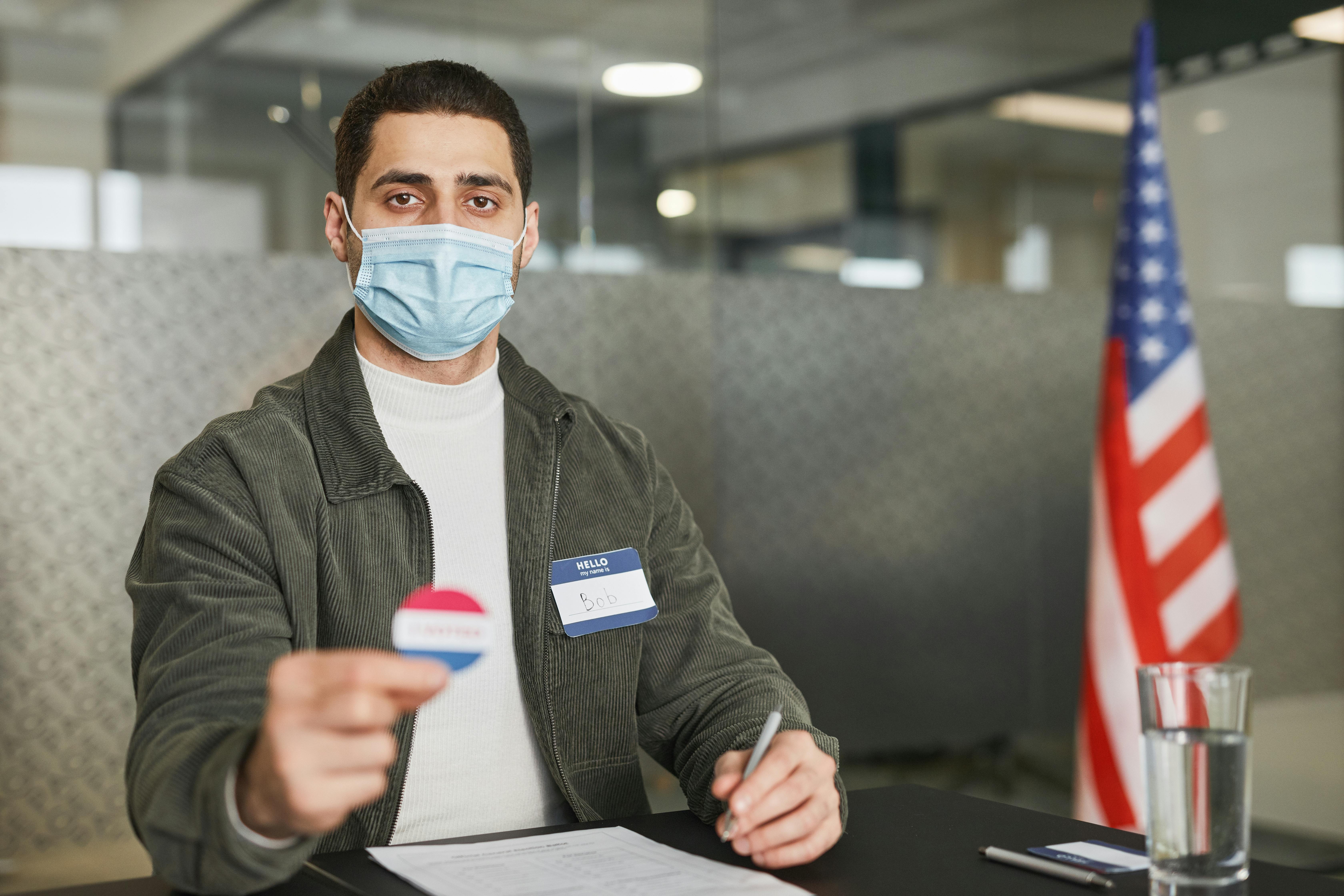 Man in office setting wearing face mask, holding voting badge with USA flag in background.