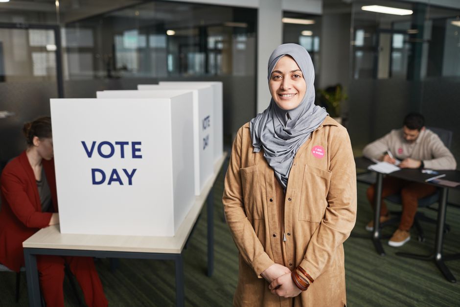 A smiling woman in a hijab stands at a voting booth on election day in a modern indoor setting.