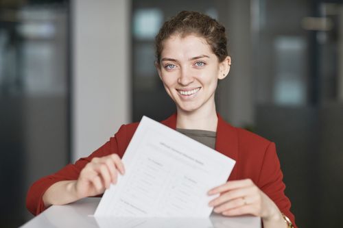 Free Smiling businesswoman in red blazer holding a document indoors, exuding confidence. Stock Photo