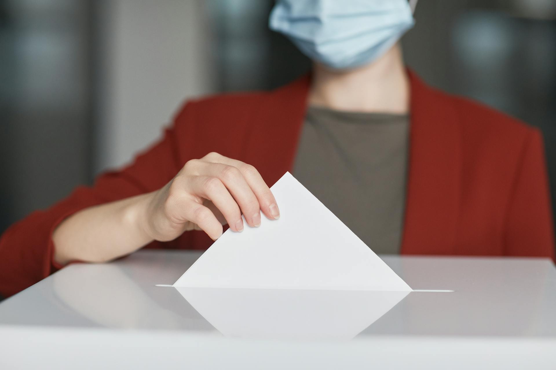 Person wearing a face mask dropping a ballot into a box to cast a vote. Emphasizes civic duty.