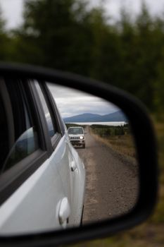 A car is reflected in the side mirror on a rural road with mountains in the distance.