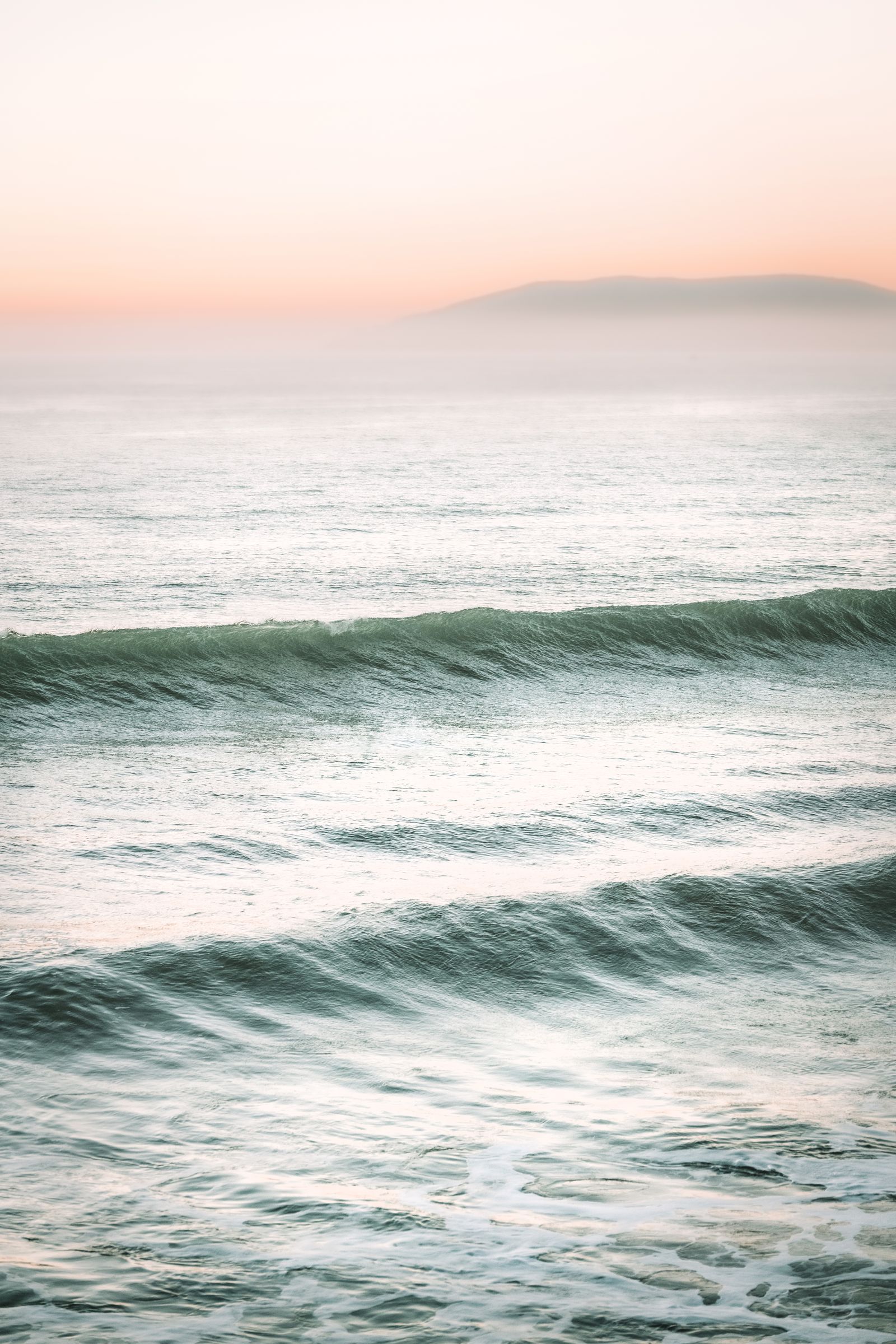 Dawn waves forming along a Pacific coastline, soft pink sky and a distant island in the mist