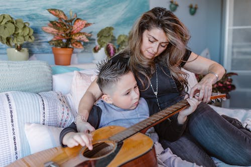 Focused little boy sitting on cozy bed and practicing acoustic guitar on bed with help of young mother during weekend at home