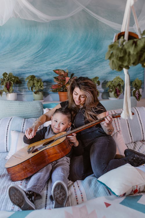 Happy young woman hugging with cute son while sitting together on comfortable bed and playing acoustic guitar during weekend at home