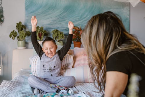 Happy boy raising arms after winning card game sitting on bed with unrecognizable mother