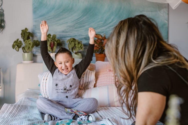 Happy Boy Raising Arms After Winning Card Game Sitting On Bed With Unrecognizable Mother
