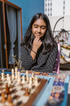 A young girl sits thoughtfully while playing chess outdoors in a bright setting.