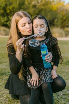 A mother and daughter share a joyful moment blowing bubbles in a park.