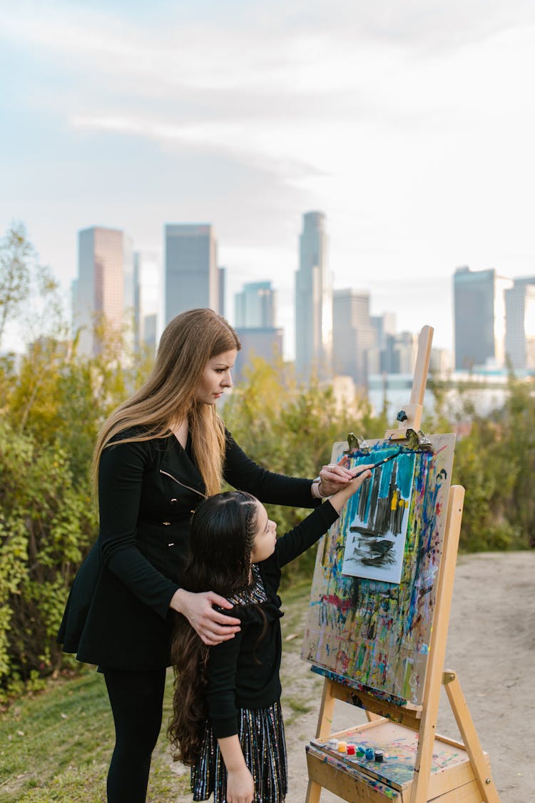 A Woman Teaching A Girl How To Paint At A Park