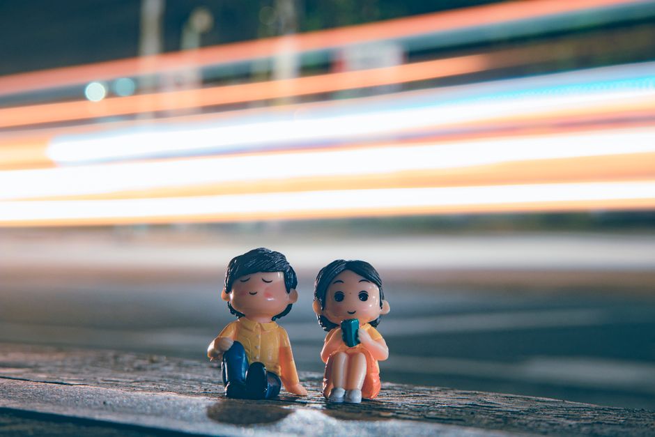 Cute figurines of boy and girl placed near asphalt road with long exposure light trails on street in evening time