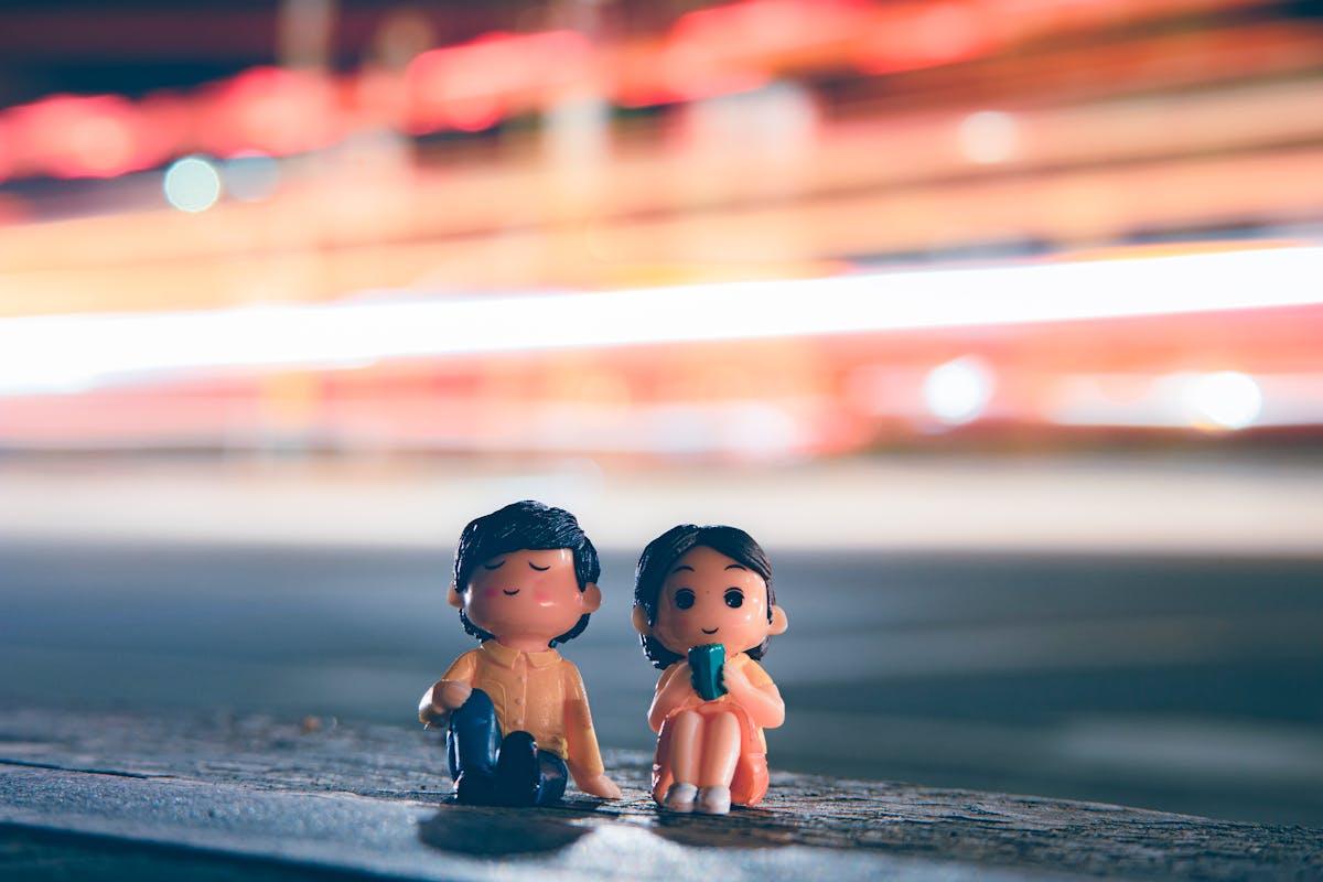 Small colorful figurines of boy and girl placed near roadside with long exposure light trails in evening time on blurred background