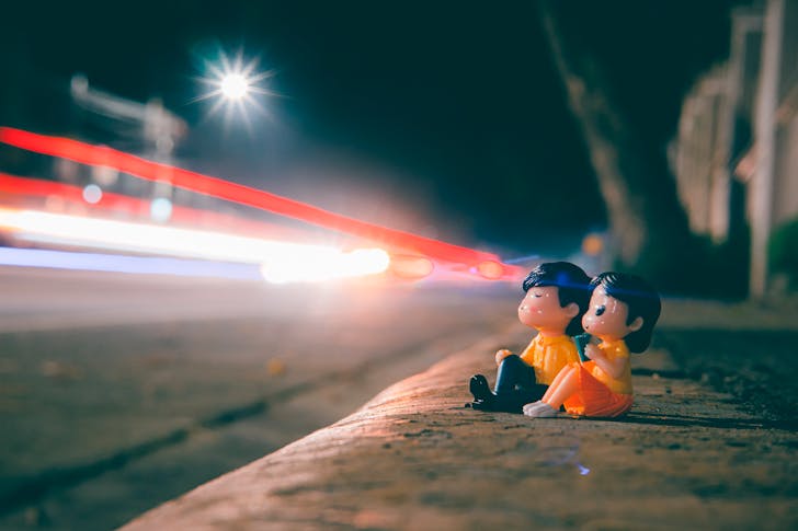 Ground level of cute couple figurines on roadside near highway with long exposure light trails in evening time on blurred background