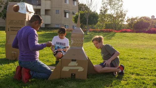Free Three boys enjoying a sunny day building a cardboard rocket in a park, showcasing creativity and teamwork. Stock Photo