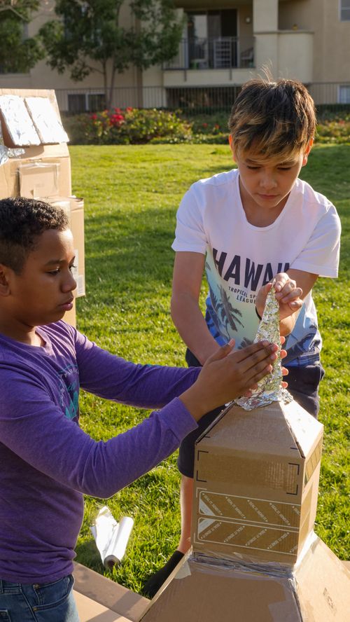 Free Two boys crafting a cardboard rocketship outdoors, enjoying a sunny day in the park. Stock Photo