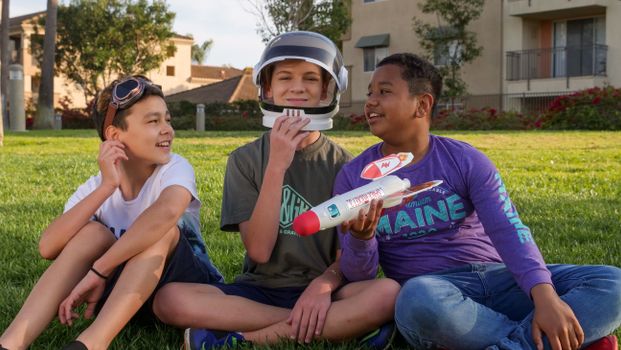 Three kids enjoy a sunny day playing with a toy rocket in a grassy field.