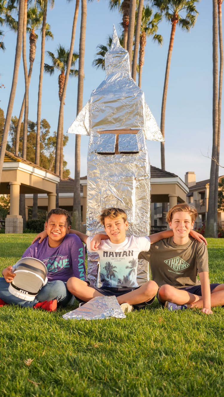 A Group Of Multiracial Boys Sitting On Green Grass With A Space Helmet