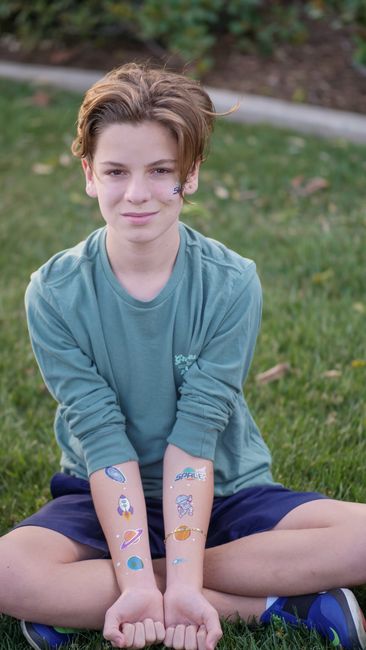 Smiling child with stickers on face and arms sitting outdoors on grass.